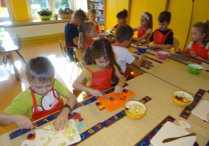 Grupa dzieci siedzi przy stole nakrytym podkładkami z rozłożonymi deskami, w ręku trzymają noże, którymi kroją owoce.